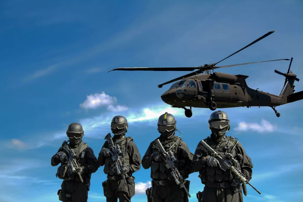 Quatre soldats en tenue de combat avec fusils, devant un hélicoptère militaire noir de type Black Hawk, sous un ciel bleu parsemé de nuages.