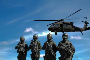 Quatre soldats en tenue de combat avec fusils, devant un hélicoptère militaire noir de type Black Hawk, sous un ciel bleu parsemé de nuages.