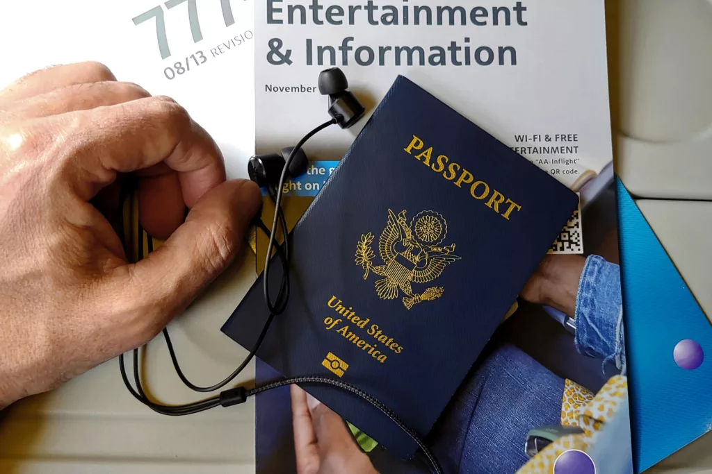 Un passeport américain bleu, des écouteurs noirs et une brochure de divertissement en vol, le tout tenu par une main.