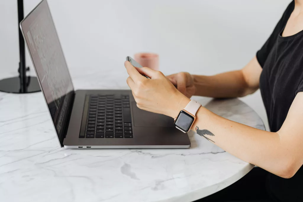 Une femme tapant sur son smartphone, un Apple Watch 40mm à son poignet, assise devant un ordinateur portable sur une table en marbre.