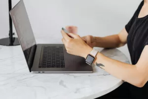 Une femme tapant sur son smartphone, un Apple Watch 40mm à son poignet, assise devant un ordinateur portable sur une table en marbre.