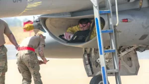 Une personne en uniforme militaire est allongée dans un compartiment d'avion, lisant un document, tandis que deux autres techniciens travaillent à proximité.