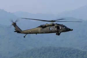 Un hélicoptère militaire de couleur sombre (type Black Hawk) en vol au-dessus de montagnes verdoyantes et brumeuses.