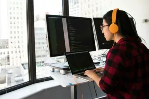 Une personne portant un casque jaune travaillant sur un ordinateur portable et deux grands écrans affichant du code.