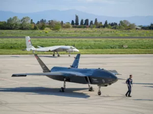 Deux drones turcs, dont un drone de combat furtif Baykar Kızılelma au premier plan et un drone d'entraînement et de reconnaissance Baykar Bayraktar Akıncı à l'arrière-plan, sont garés sur un tarmac d'aéroport. Un pilote en combinaison de vol marche près du Kızılelma.