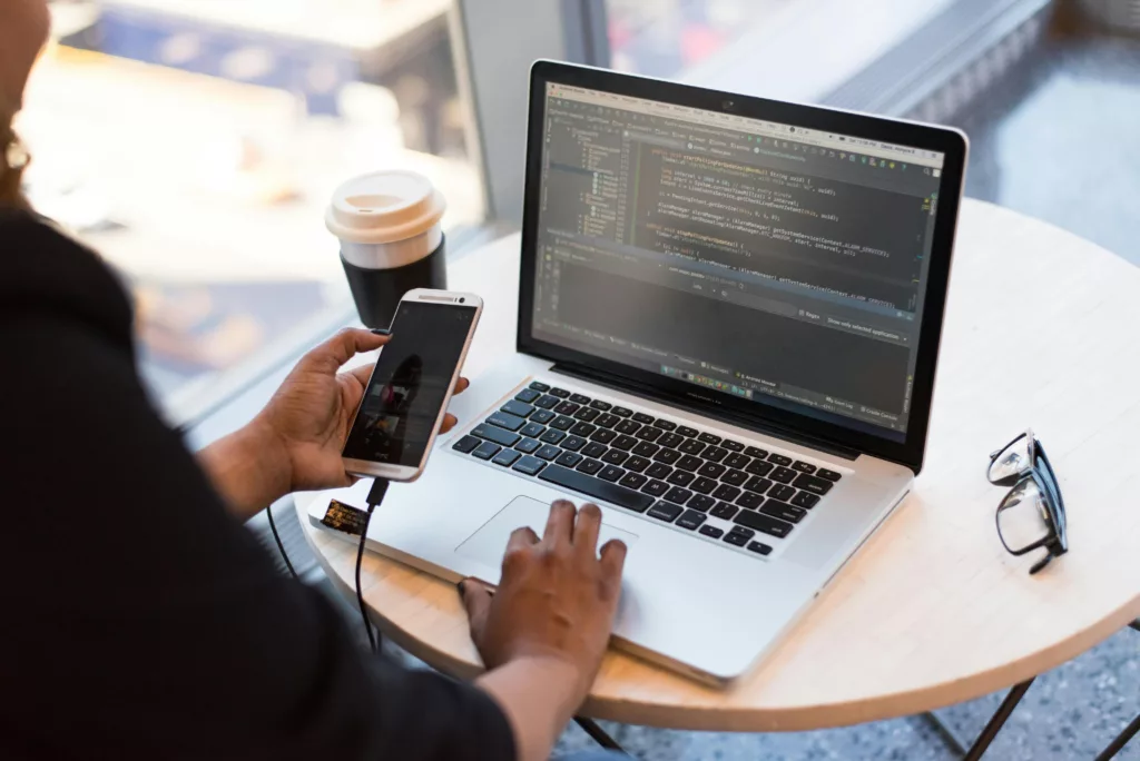 Femme développeuse utilisant un ordinateur portable et un smartphone, avec du code affiché à l'écran, dans un café.