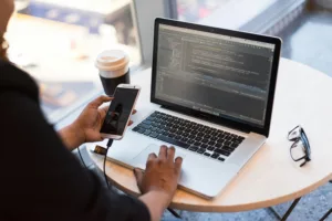 Femme développeuse utilisant un ordinateur portable et un smartphone, avec du code affiché à l'écran, dans un café.
