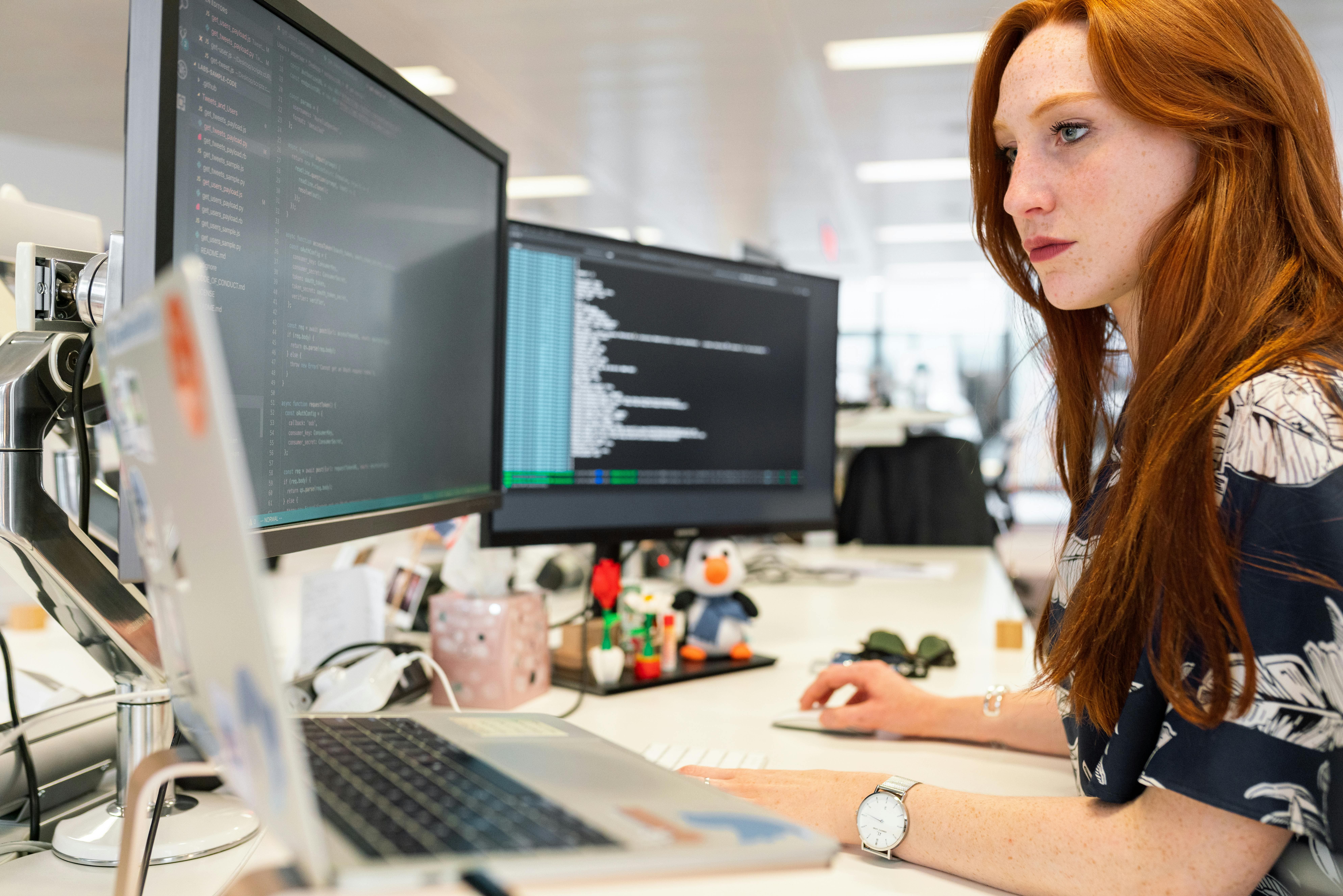 Femme rousse travaillant sur deux écrans d'ordinateur affichant du code.