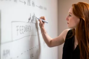 Une personne aux cheveux roux écrit sur un tableau blanc, penchée vers l'avant.
