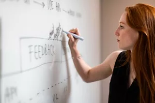 Une personne aux cheveux roux écrit sur un tableau blanc, penchée vers l'avant.